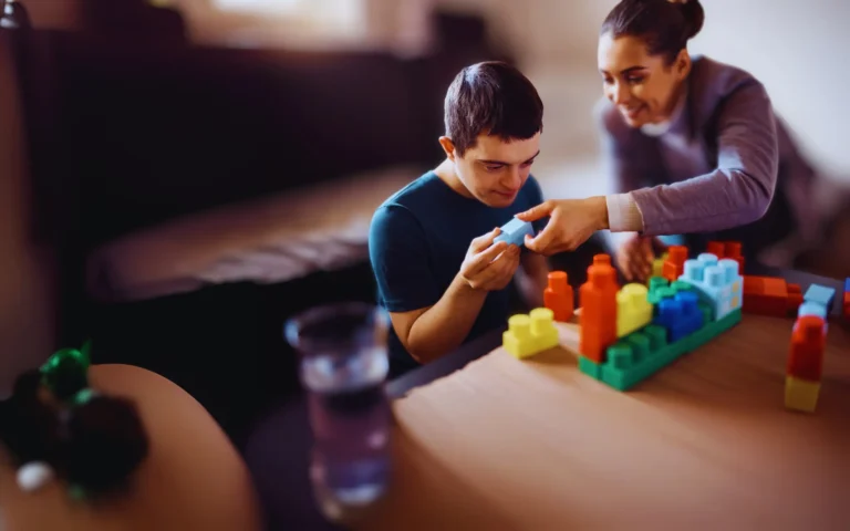 A young man with a learning disability sits at a table building with colourful blocks, supported by a smiling caregiver who gently guides him.