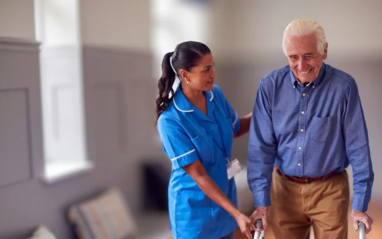 caregiver in a blue uniform supports an older man using a walking frame