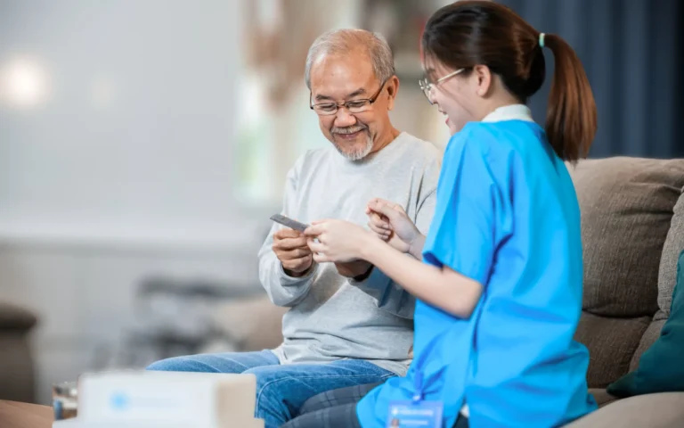 Caregiver in blue scrubs sharing a warm moment on a sofa with an elderly man in his home, both smiling