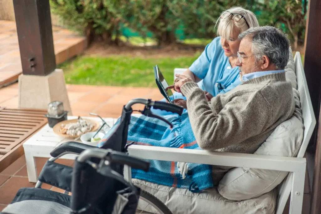 A Careline 24 carer in West Sussex compassionately assists an elderly gentleman using a tablet computer in a sunny garden, enabling social connection and digital inclusion.