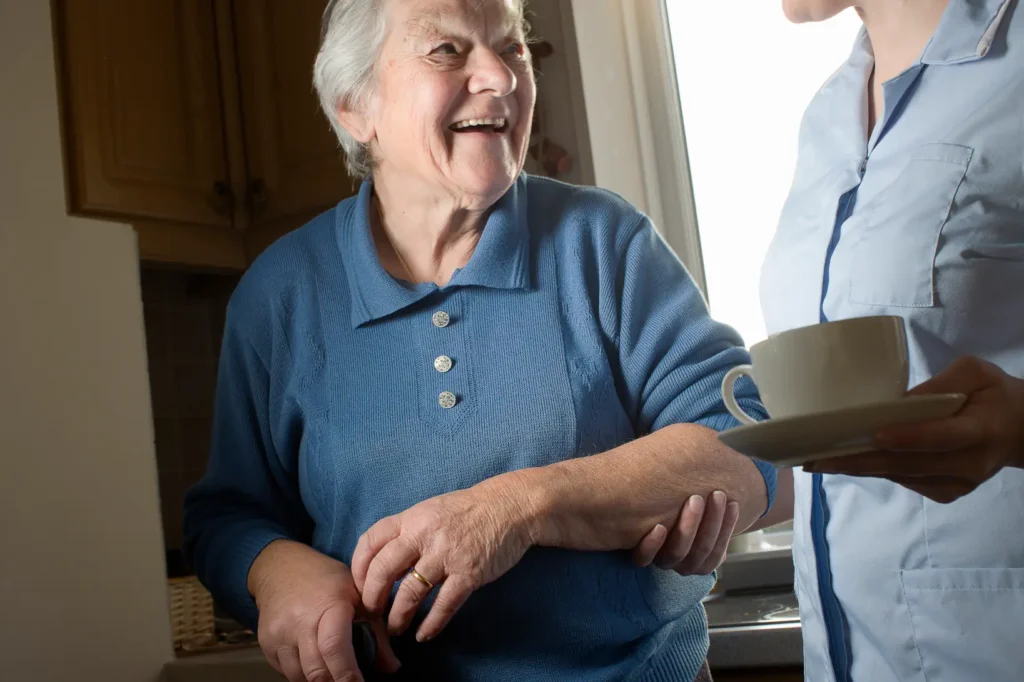 A friendly, professional carer carefully carries a warm cup of tea to an elderly lady relaxing comfortably in her own living room.