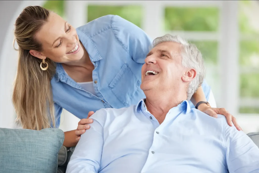 A cheerful Careline 24 carer shares a happy, conversational visit with an elderly gentleman on the sofa in his Bognor Regis home.