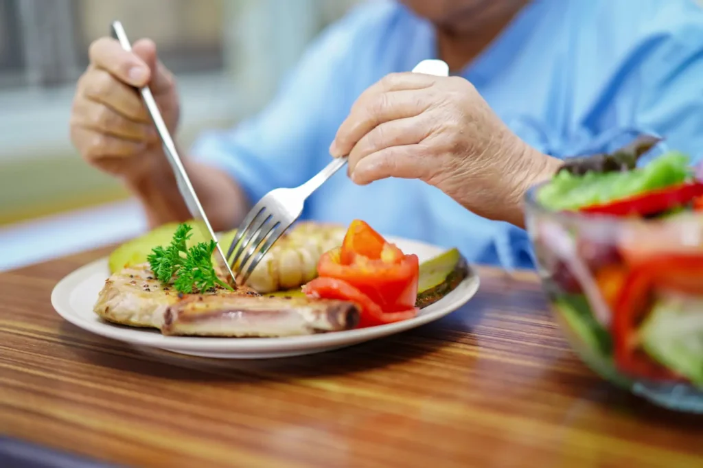 A compassionate carer provides supportive mealtime assistance as an elderly Asian lady enjoys a nutritious breakfast in her own dining room.