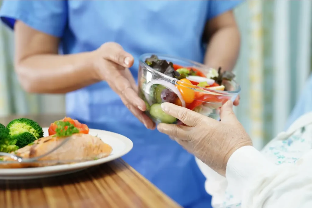 A compassionate carer provides supportive mealtime assistance to an elderly Asian lady enjoying breakfast in her own home.