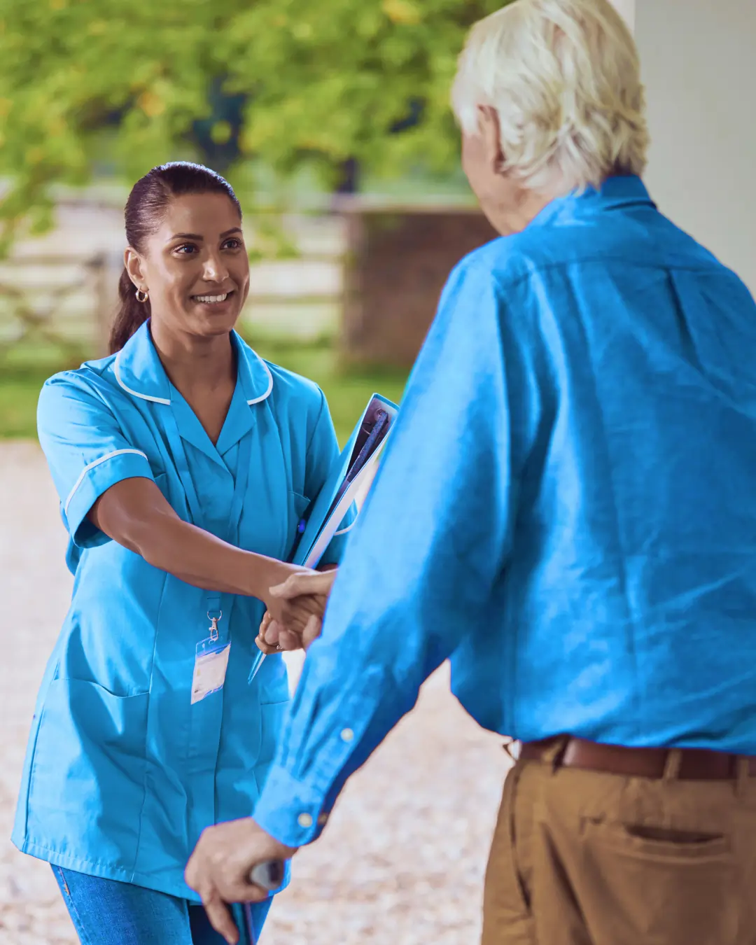caregiver in a blue uniform smiles warmly as she shakes hands with an elderly man outside his home