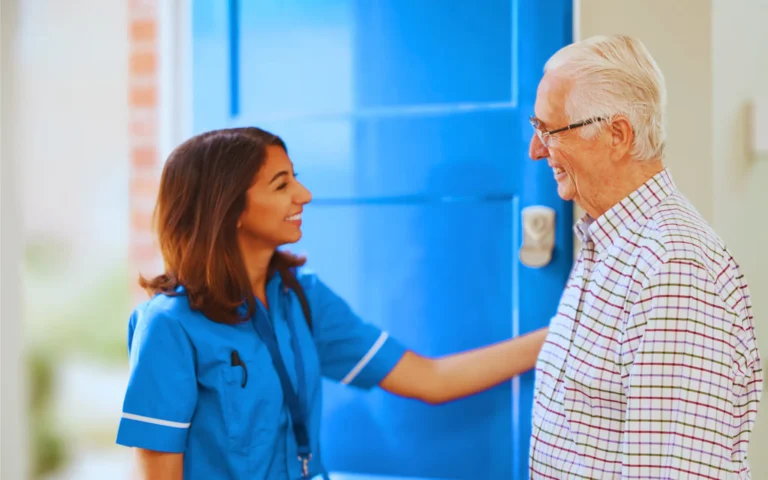 caregiver in a blue uniform greets an elderly man at his front door